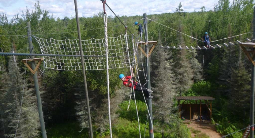 A person wearing safety gear is attached to ropes as they navigate a ropes course in a wooded area. 